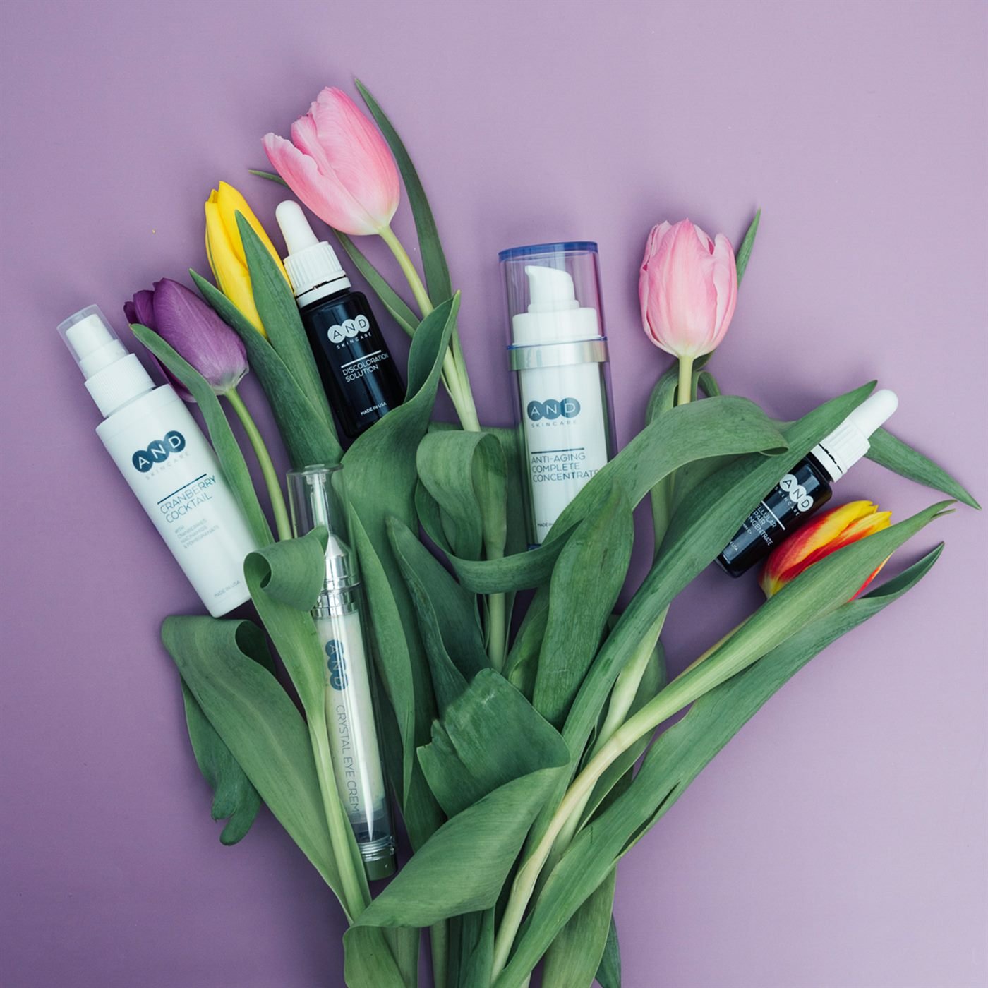 AND skincare Produkte zwischen frischen Tulpen auf fliederfarbenem Hintergrund